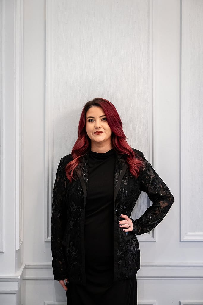 Young Women with red hair standing in front of a white wall with crown molding wearing a black blazer adorned with lace and sequence and black dress. J. Parsons Photography