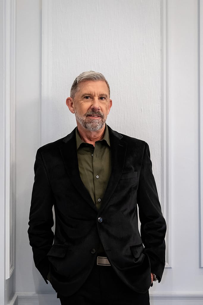 Older Gentleman standing in front of a white wall with crown molding earring a black velvet jacket and an olive green shirt. J. Parsons Photography
